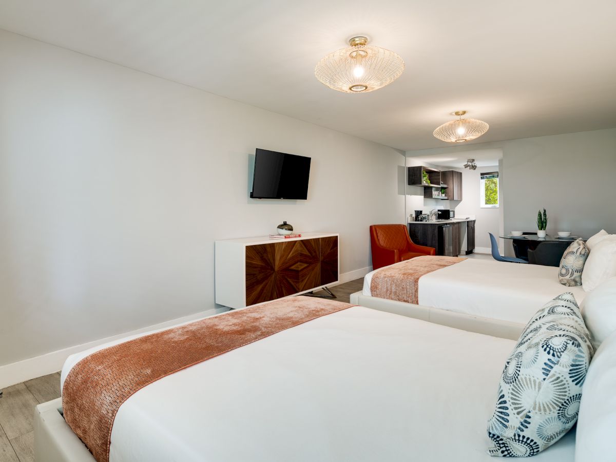 A bright, modern hotel room with two queen beds, a wall-mounted TV, a small dresser, and an open-plan kitchenette area in the background.