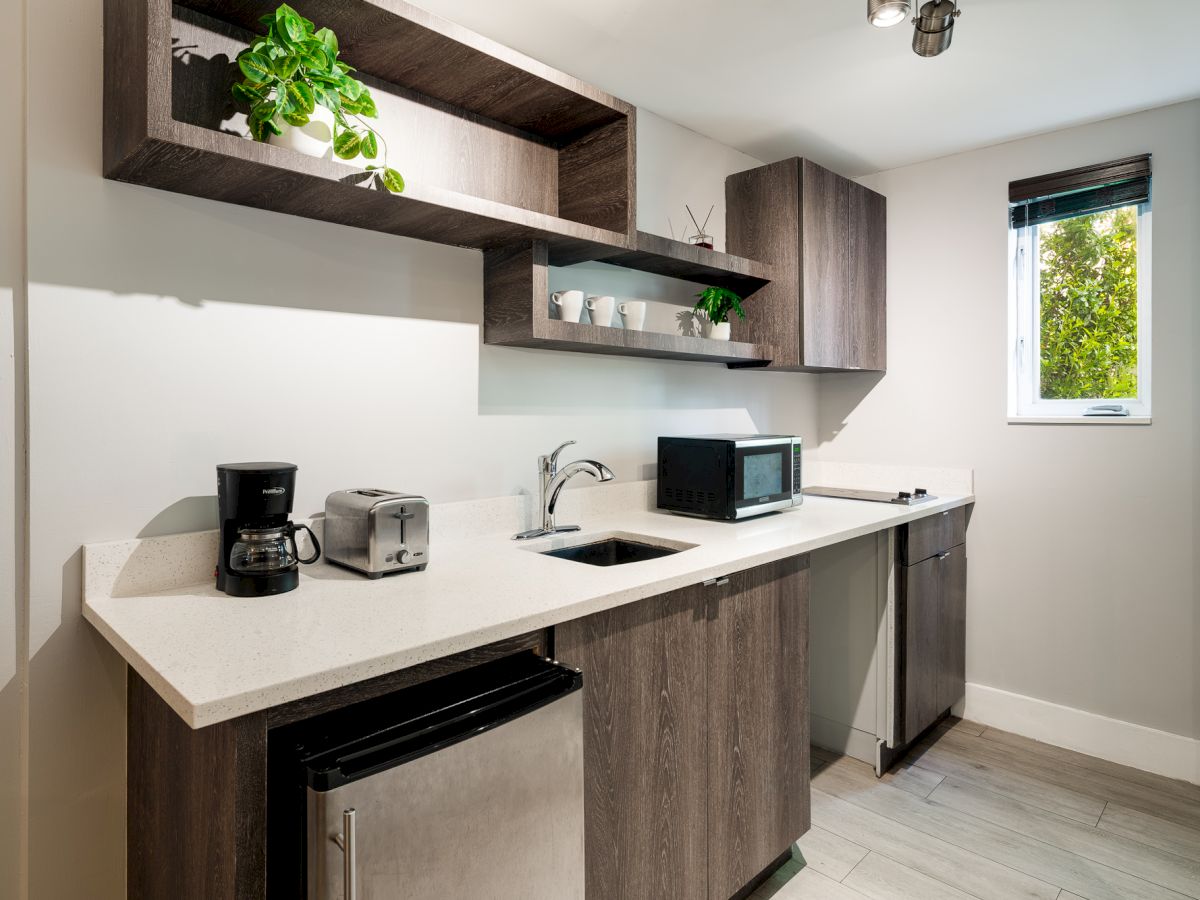 A compact modern kitchenette with dark wood cabinets, white countertop, a coffee maker, toaster, microwave, sink, small fridge, and a window near the end.