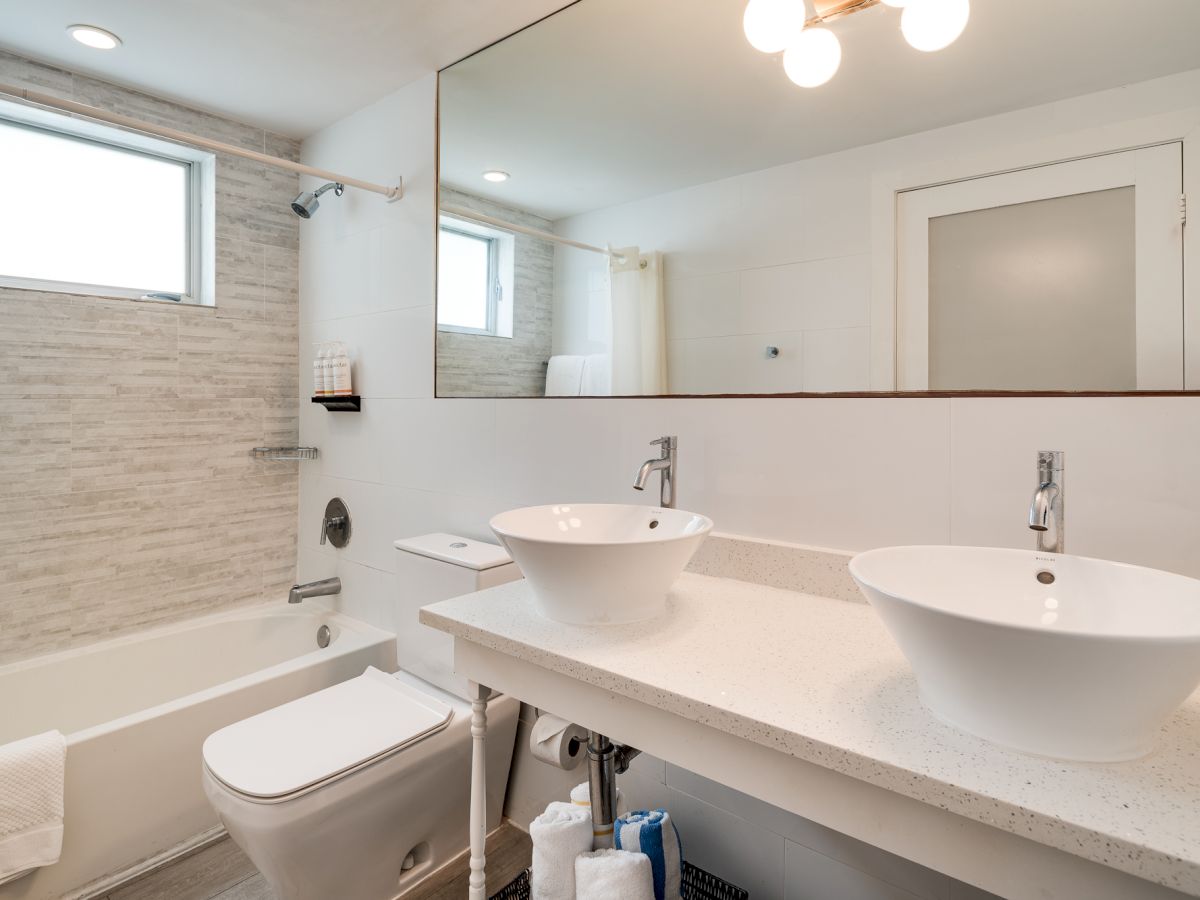 A modern bathroom with a double vanity, round vessel sinks, a toilet, and a bathtub/shower combo with a frosted window. Top it at 140 characters, always ending the sentence.