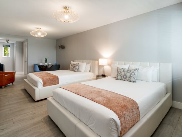A modern hotel room with two white upholstered beds, soft peach throws, neutral walls, a nightstand lamp, and a seating area in the background.