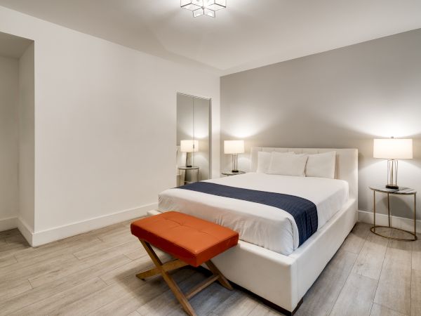 A modern hotel bedroom with a white upholstered bed, two nightstands, lamps, and a small orange bench at the foot of the bed, neutral decor.
