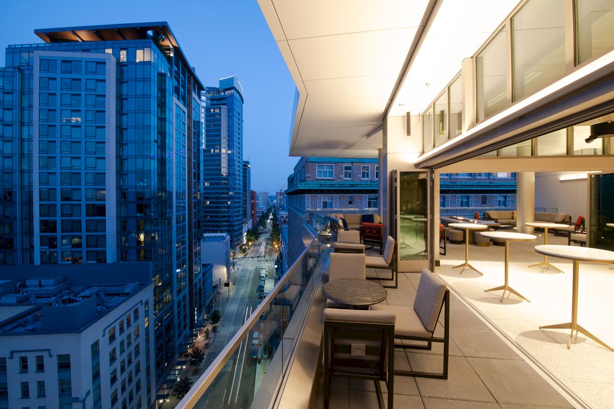 A modern balcony with tables and chairs overlooks a cityscape with tall buildings during dusk.
