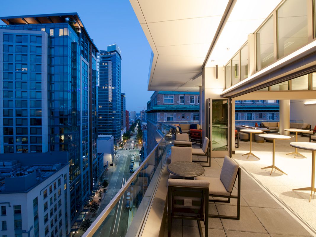 A modern balcony with tables and chairs overlooks a cityscape with tall buildings during dusk.
