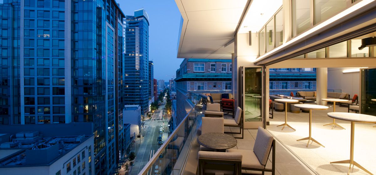 A modern balcony with tables and chairs overlooks a cityscape with tall buildings during dusk.