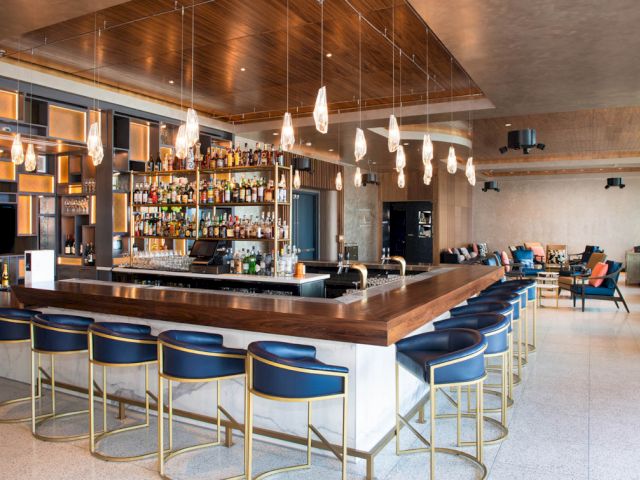 A wide view of a bar with blue stools, a wood counter, and shelves of bottles.