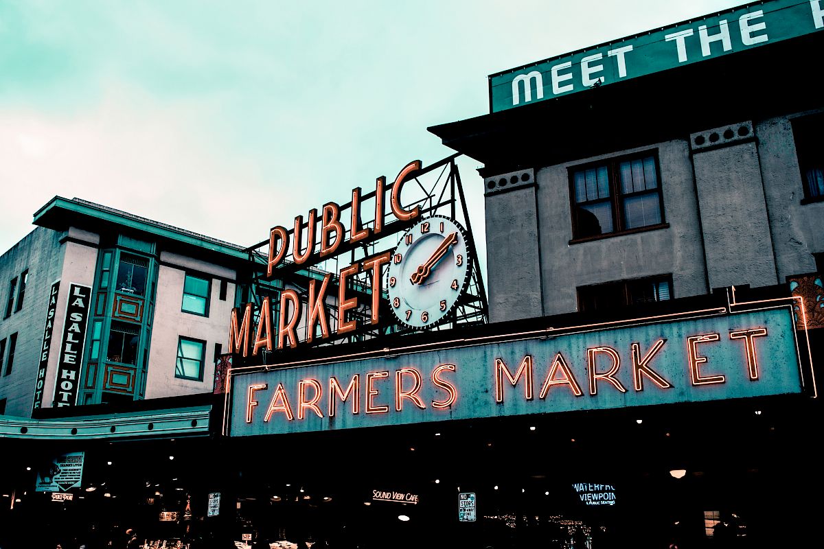 The image shows a public market with neon signs for "Public Market" and "Farmers Market" against a building facade.
