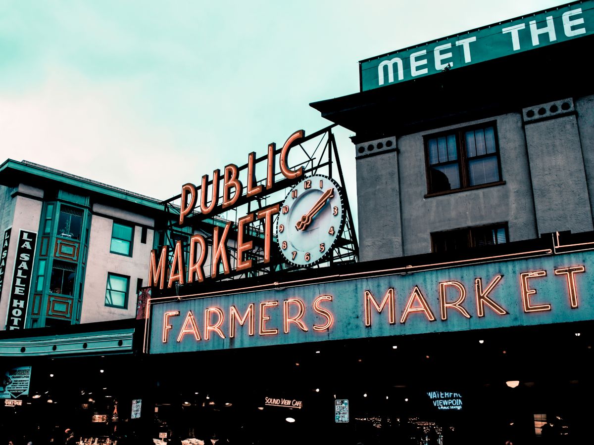 The image shows a public market with neon signs for "Public Market" and "Farmers Market" against a building facade.