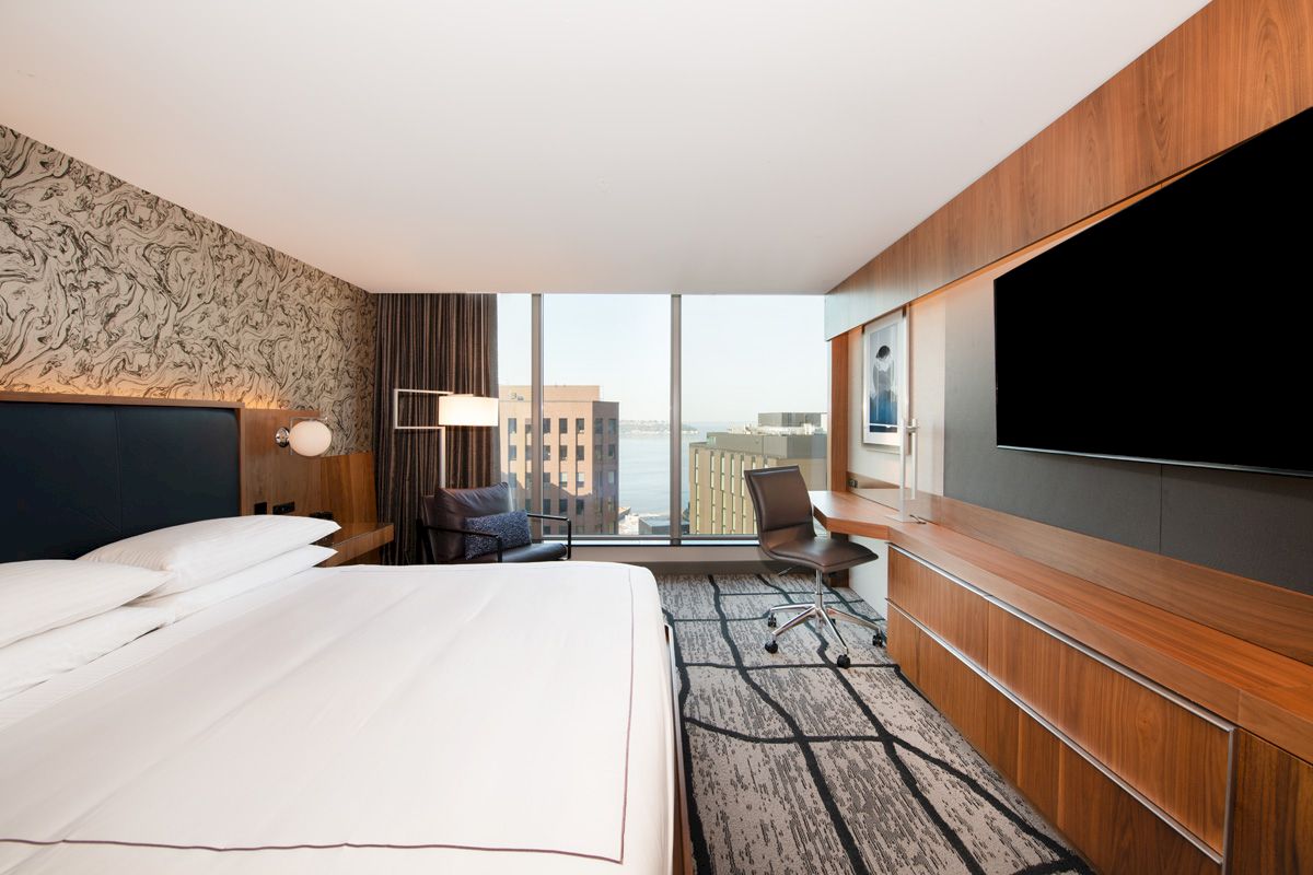 A modern hotel room with a large bed, desk, TV, and chair, featuring a city view through the window.