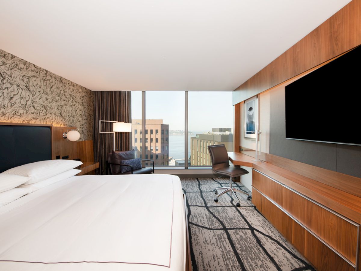 A modern hotel room with a large bed, desk, TV, and chair, featuring a city view through the window.