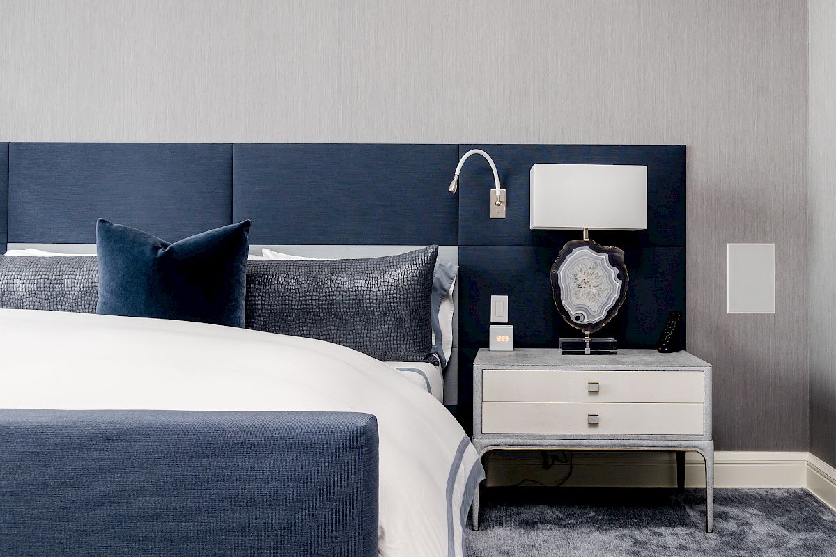 A modern bedroom with a blue upholstered bed, decorative pillows, a white nightstand with a lamp, and a textured gray wall.