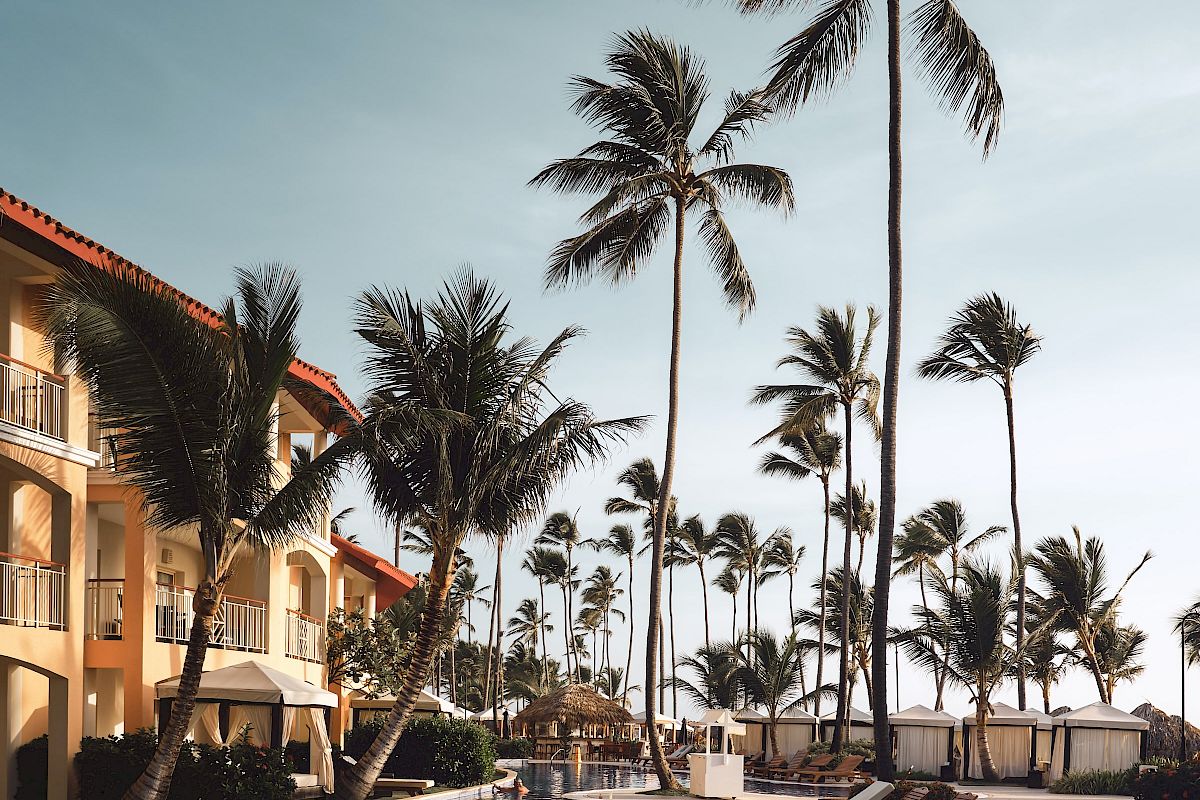 A tropical resort scene with palm trees, a pool, lounge chairs, and a building under a clear sky.
