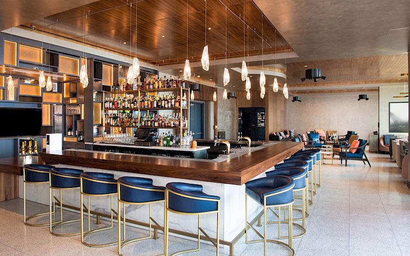 A modern bar with a wooden counter, stylish bar stools, hanging lights, and a well-stocked liquor shelf in a chic, open lounge area.