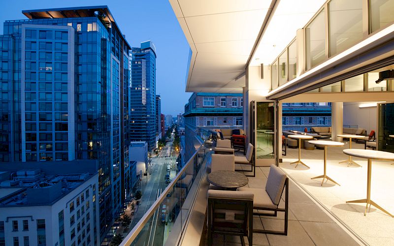 A modern rooftop terrace overlooking a city street, with seating and tables, surrounded by tall buildings in the evening.