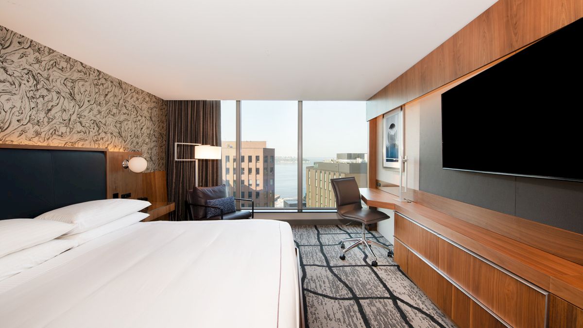 A modern hotel room with a large bed, wall-mounted TV, desk, chair, and window view of buildings.